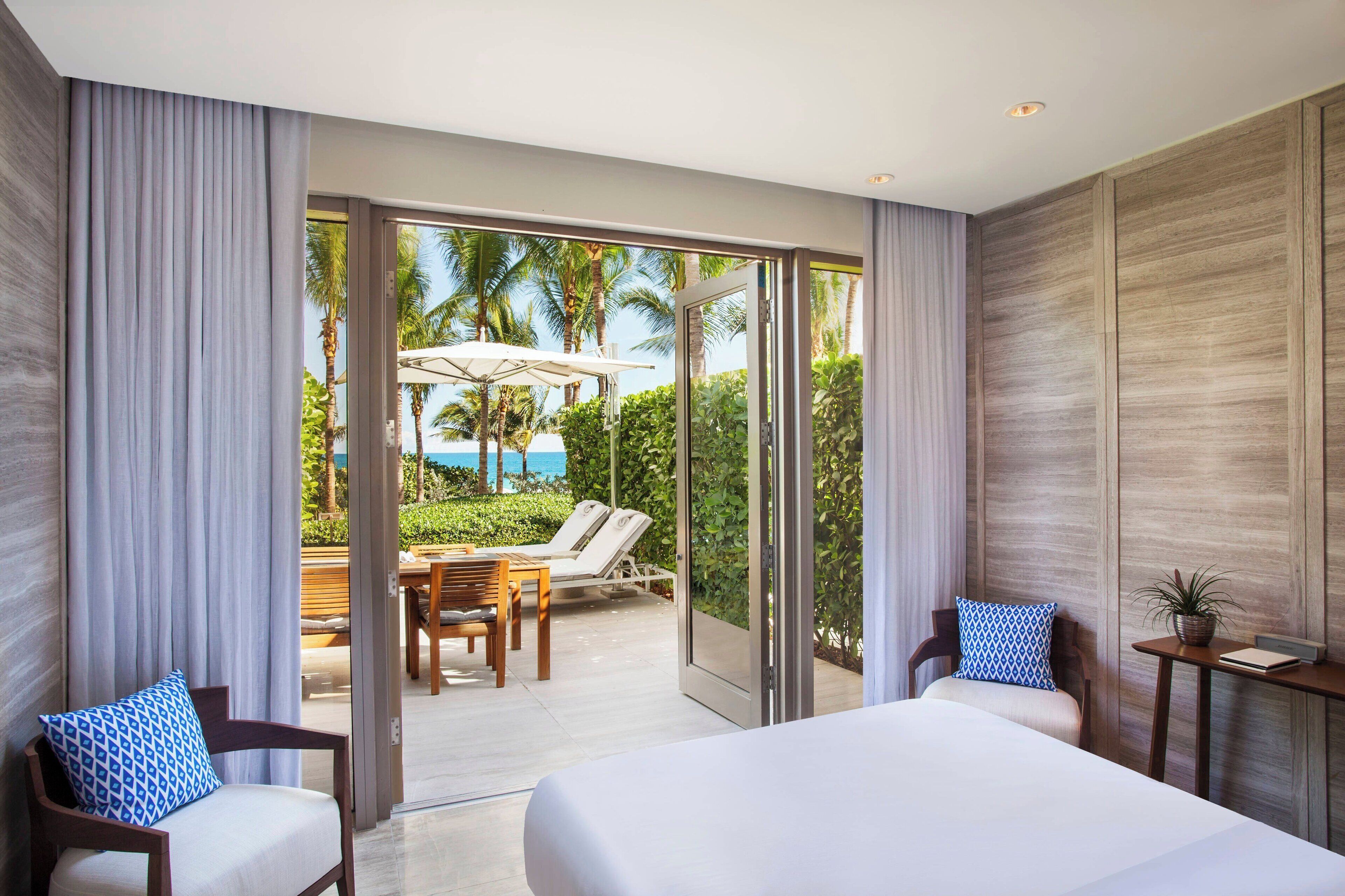 A modern hotel room with a large bed, two chairs, a small table, and a view of a pool and palm trees.