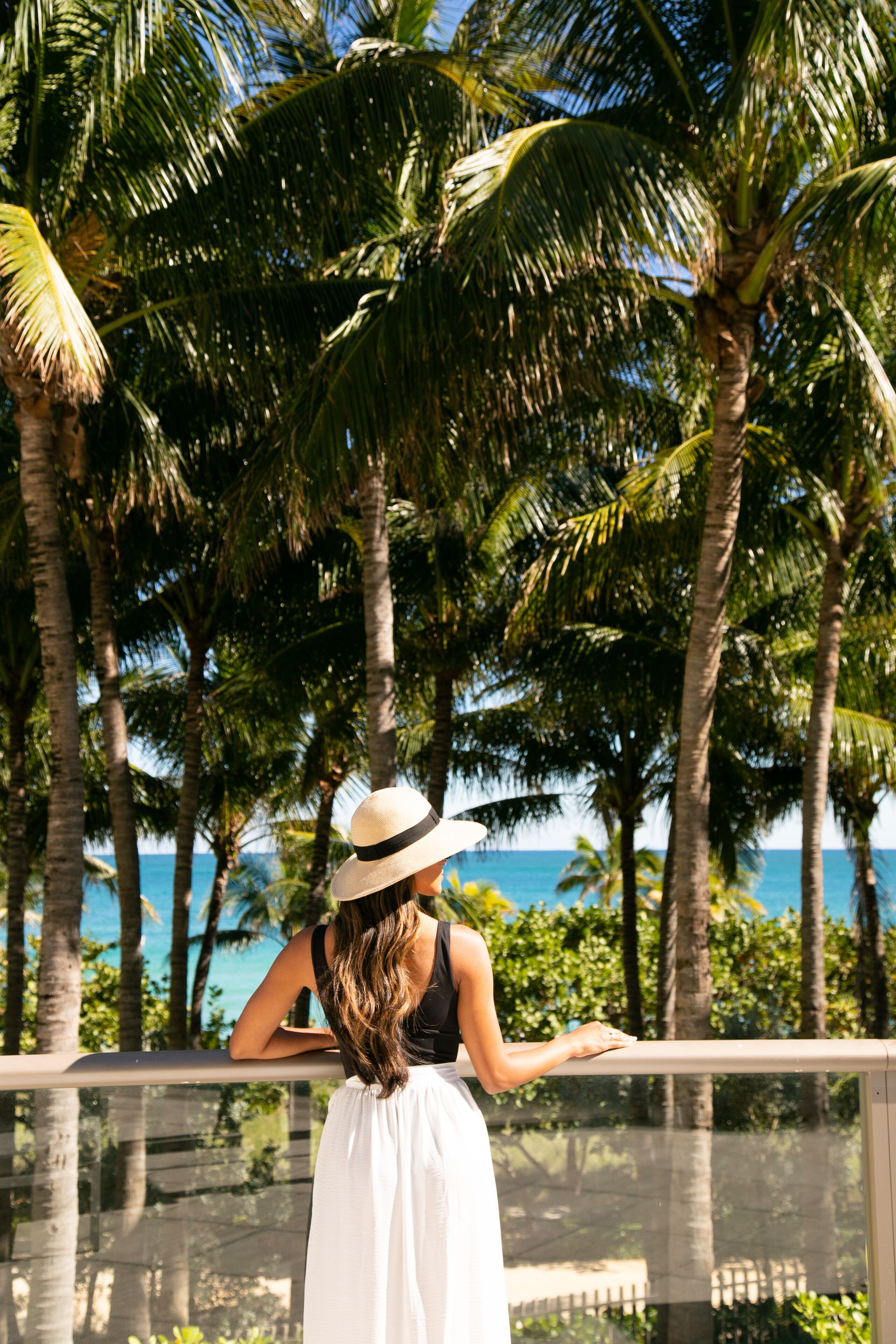 Una mujer con vestido y sombrero blancos, de pie junto a la barandilla de un balcón, con vista a una playa bordeada de palmeras y el océano.