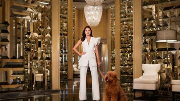 Une femme vêtue d’un tailleur blanc se tient dans un hall luxueux, accompagnée d’un chien. Elle est entourée de panneaux muraux géométriques réfléchissants et d’un lustre.