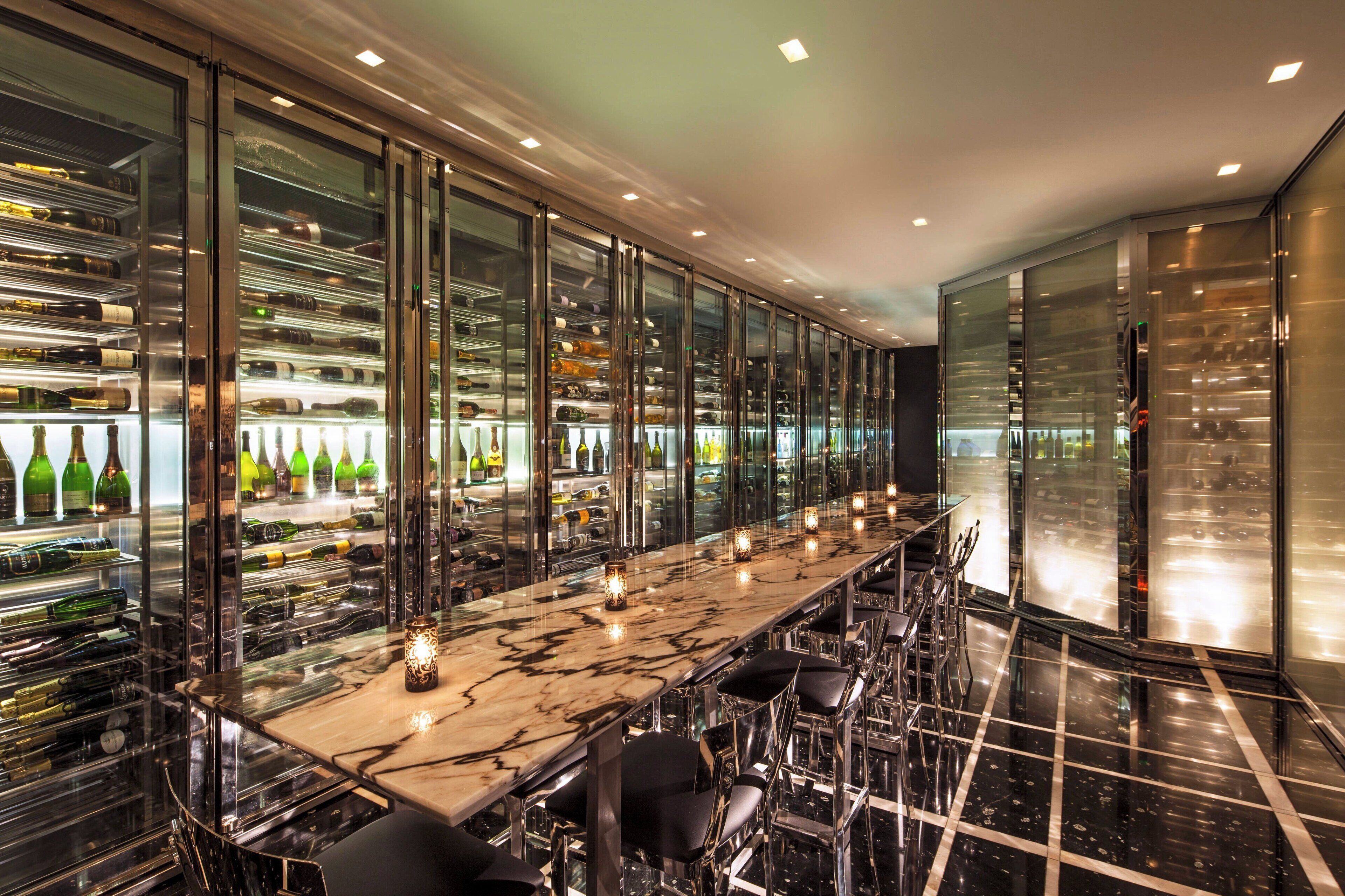 A long bar with a marble countertop and black chairs, surrounded by glass walls displaying wine bottles.
