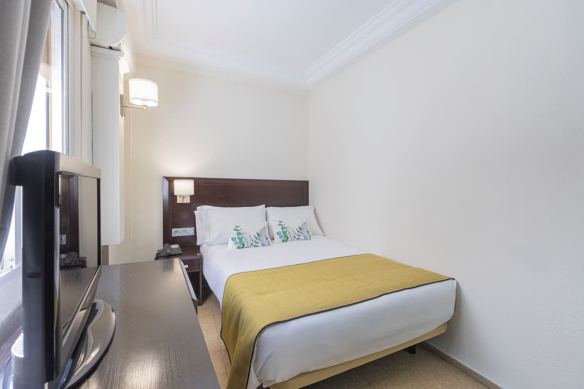 Una habitación de hotel con cama, televisión sobre un mueble, mesita de noche y velador.