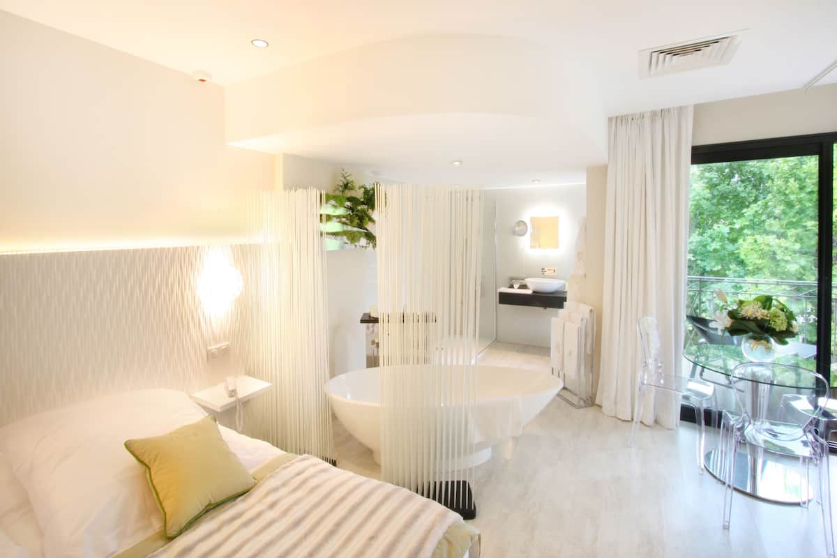 Une chambre d’hôtel moderne dotée d’un grand lit, d’une salle de bain séparée avec baignoire, et d’un coin repas avec une table et des chaises en verre.