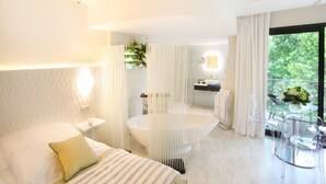 Una habitación de hotel moderna con una cama grande, un baño independiente con bañera y una zona de comedor con mesa y sillas de vidrio.