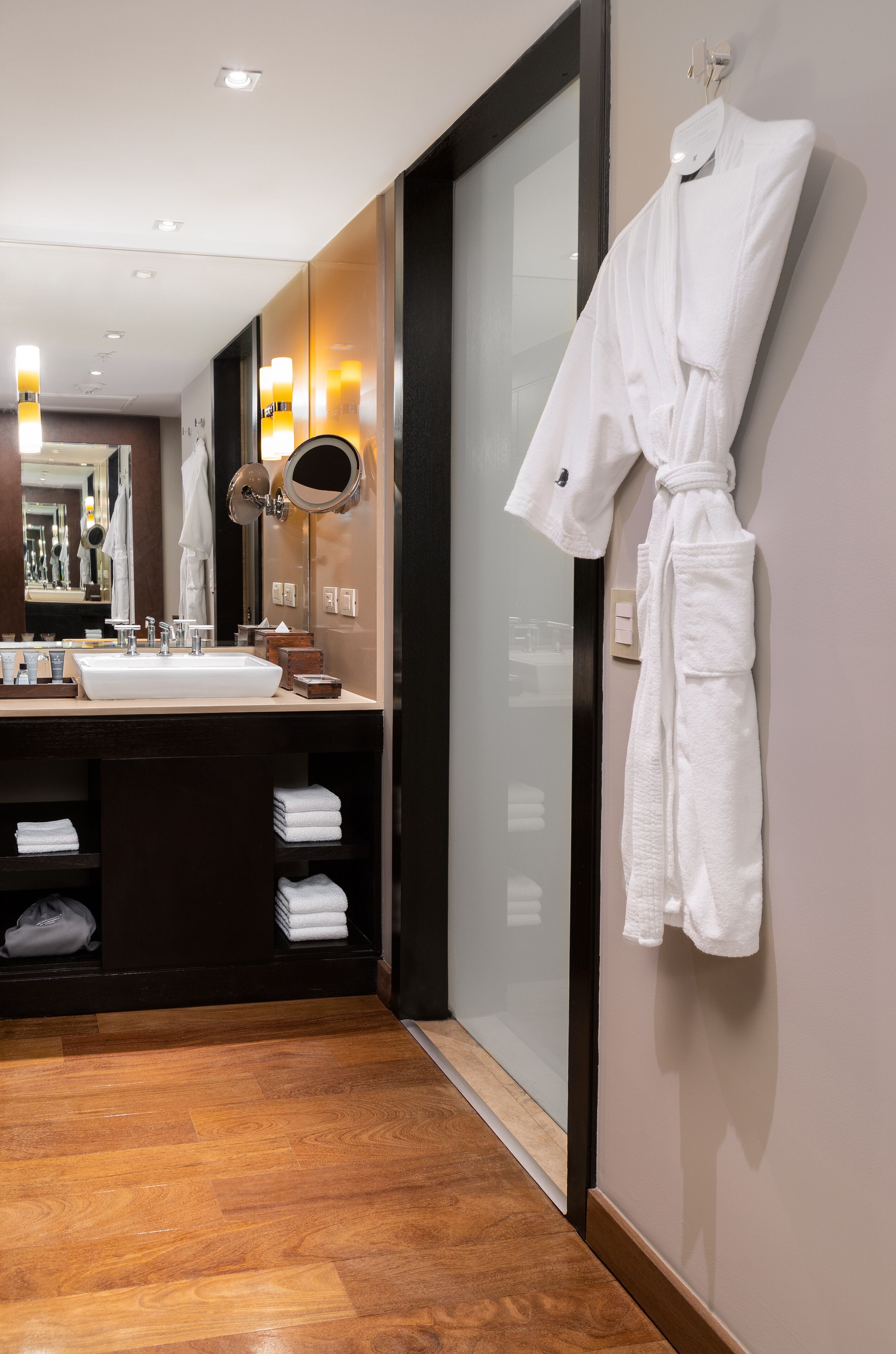 A modern bathroom with a large mirror, a white sink, and a robe hanging on the wall.
