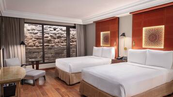 Habitación de hotel con dos camas, una mesita pequeña y una silla. Se ve una pared de piedra a través de la ventana, y paneles decorativos en la pared.