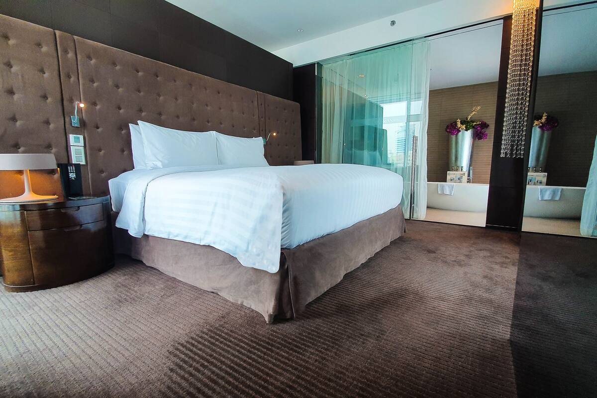 Ein modernes Hotelzimmer mit einem großen Bett, einem Nachttisch mit Lampe, einer Glasdusche und Blick auf die Stadt.