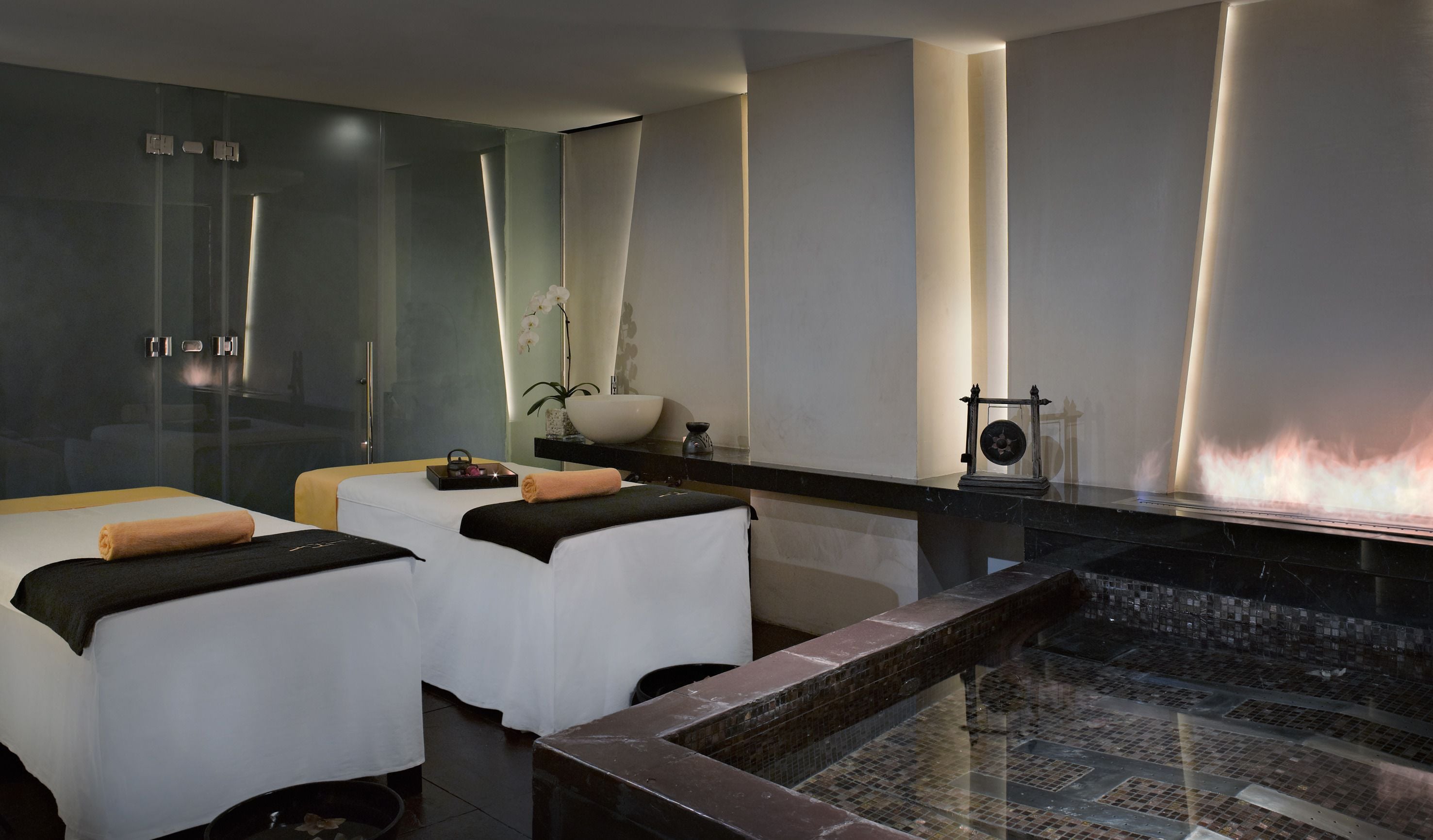 A modern spa room with two massage tables, a large mirror, a wall-mounted fireplace, and a small sink area.