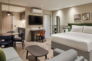 A hotel room with a bed, a desk with a chair, a dining table with chairs, a TV, and artwork on the wall.