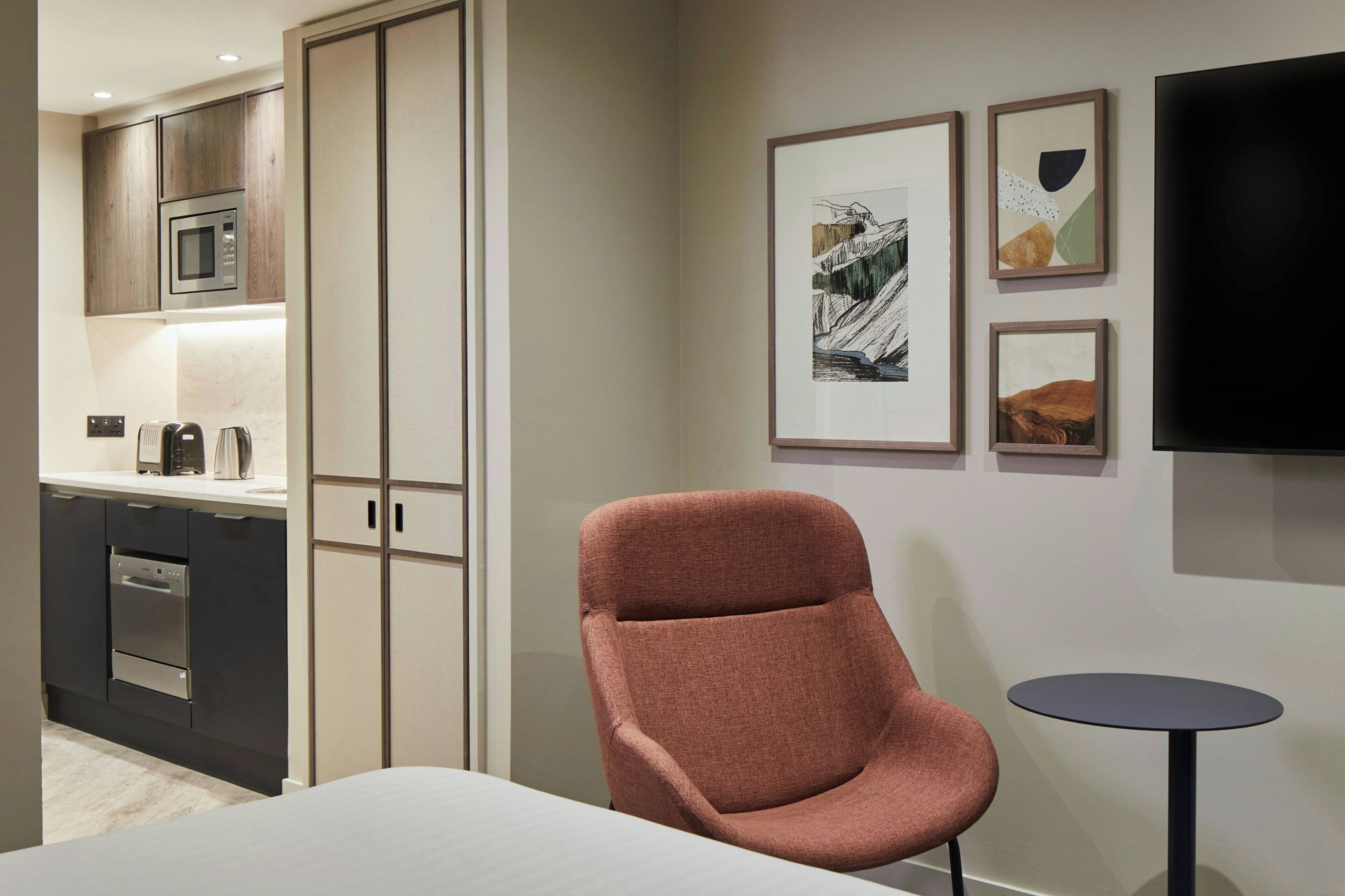 Habitación de hotel moderna con cama, sillón, pequeña mesa, televisor y kitchenette.