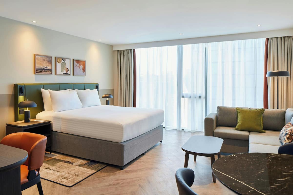 A hotel room with a bed, a sofa, a small table, and a chair. There is a window with curtains and artwork on the wall.