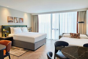 A hotel room with a large bed, a desk with a chair, a small table, and a view of the city through large windows.