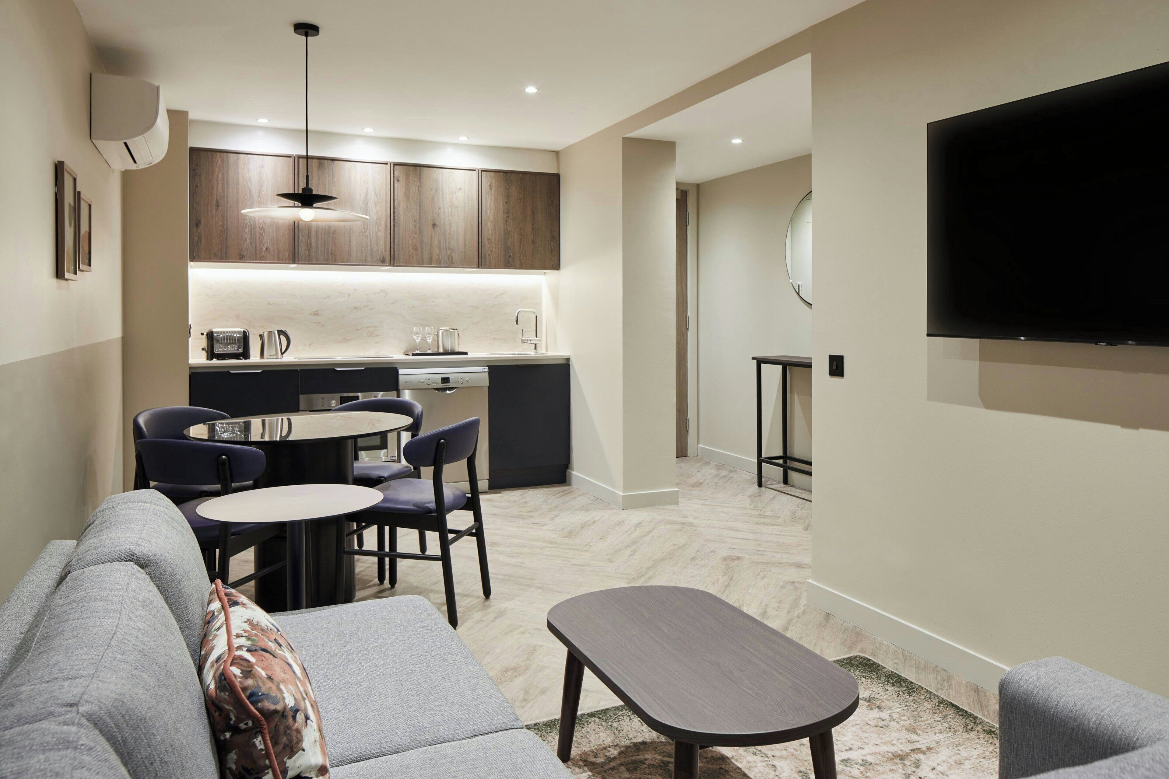 A modern living room with a grey sofa, a small round table, and a flat-screen TV mounted on the wall.