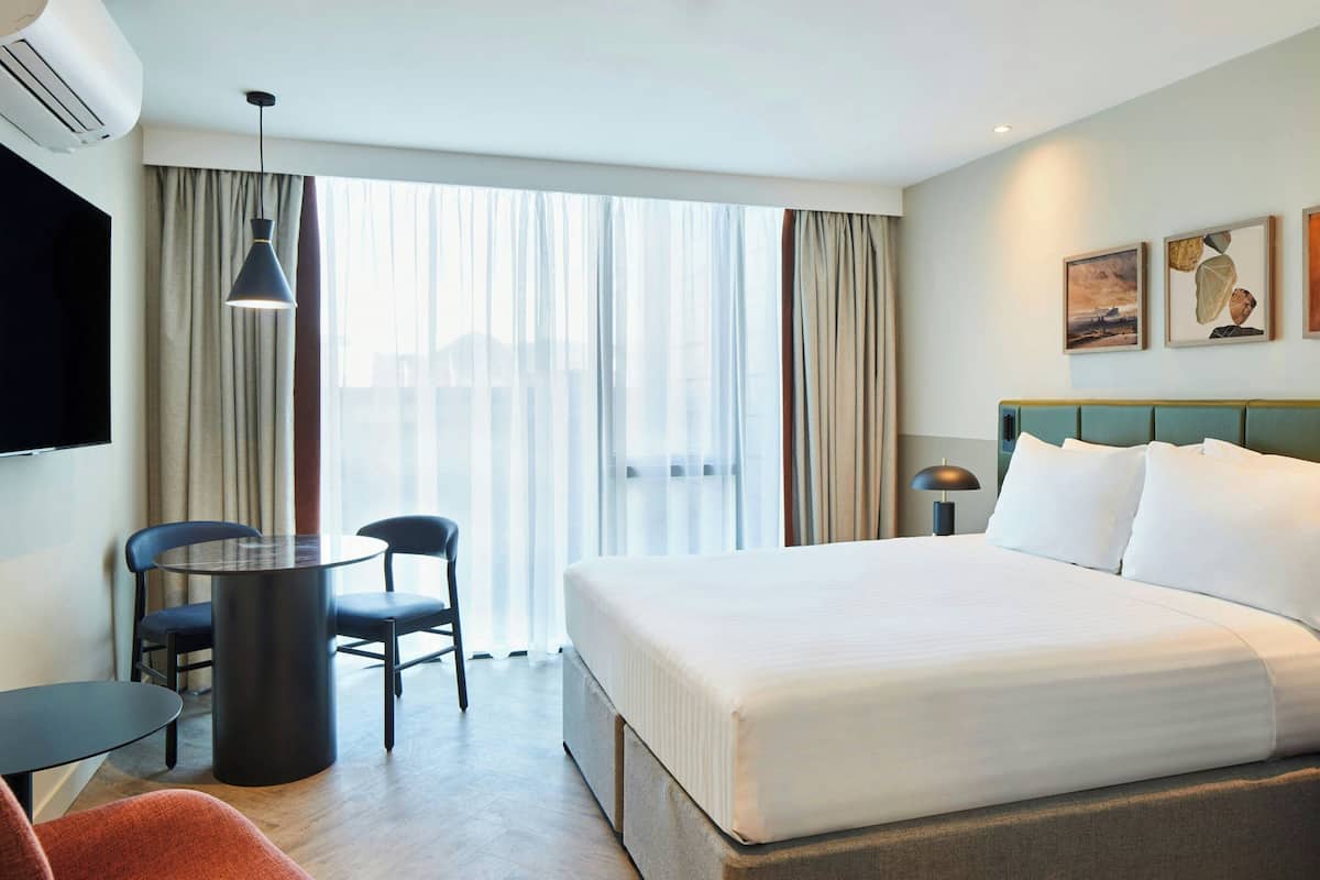 A hotel room with a bed, a desk, two chairs, a TV, and a window with curtains.