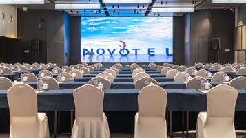 A conference room with rows of tables and chairs, a large screen displaying 'NOVOTEL,' and a staircase in the background.