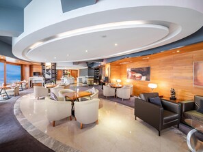 A modern hotel lobby with a curved ceiling, seating areas, a bar, and a large window offering a view.
