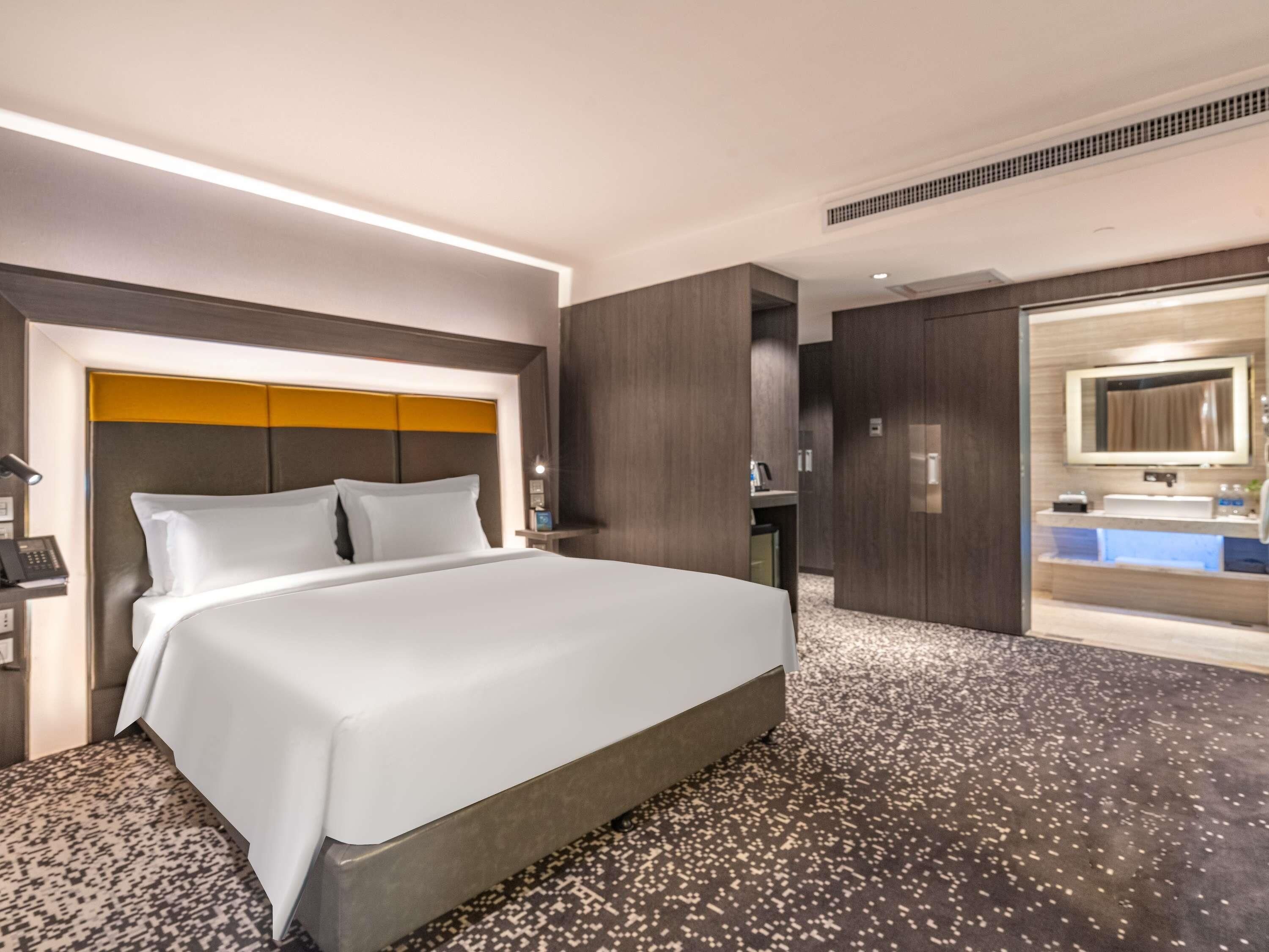 Une chambre d’hôtel moderne dotée d’un grand lit, d’un bureau intégré, d’une salle de bain avec un lavabo et un miroir, et d’un sol recouvert de moquette.