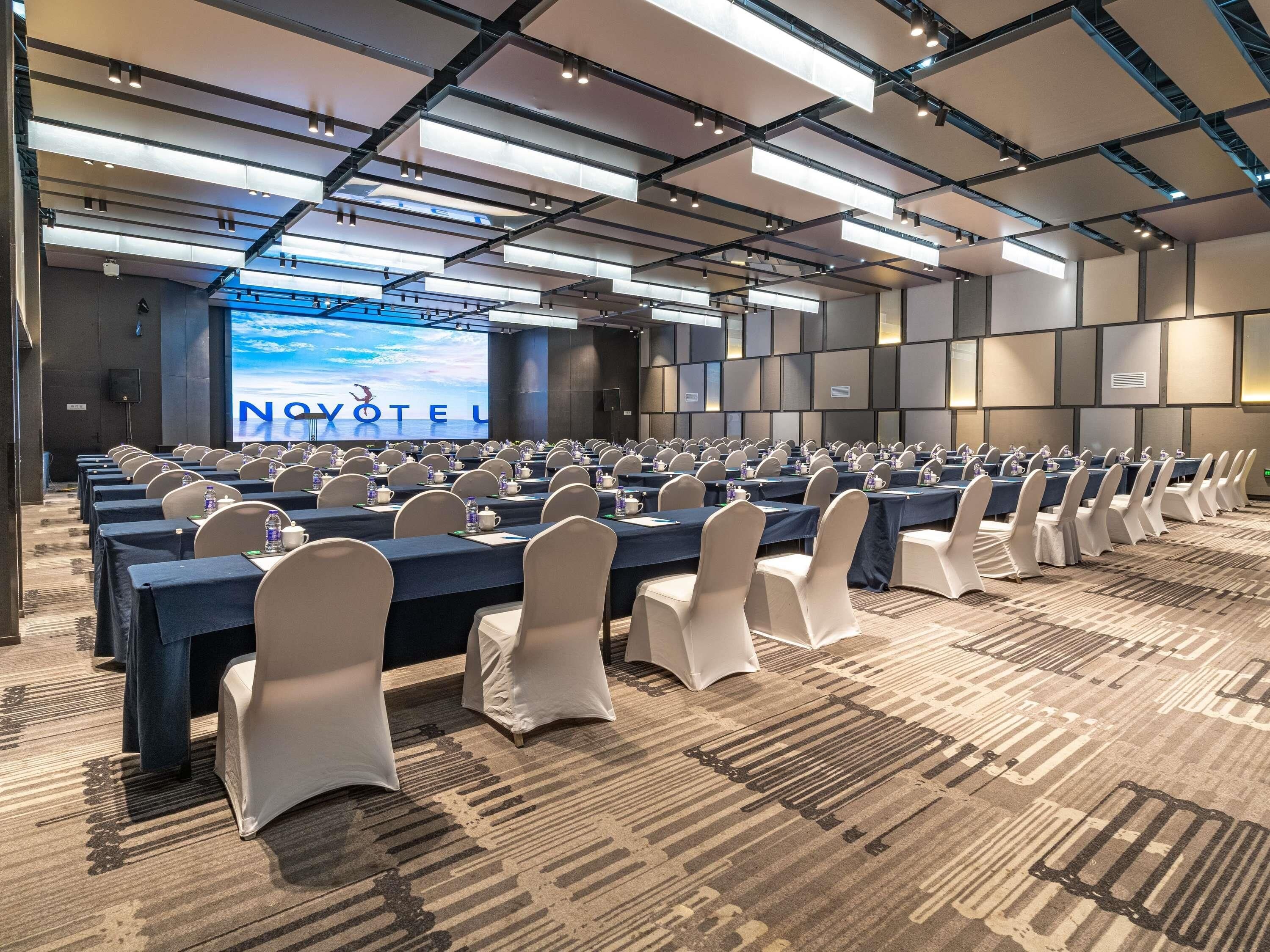 A conference room with rows of tables and chairs, a large screen displaying 'NOVOTEL,' and a modern ceiling design.