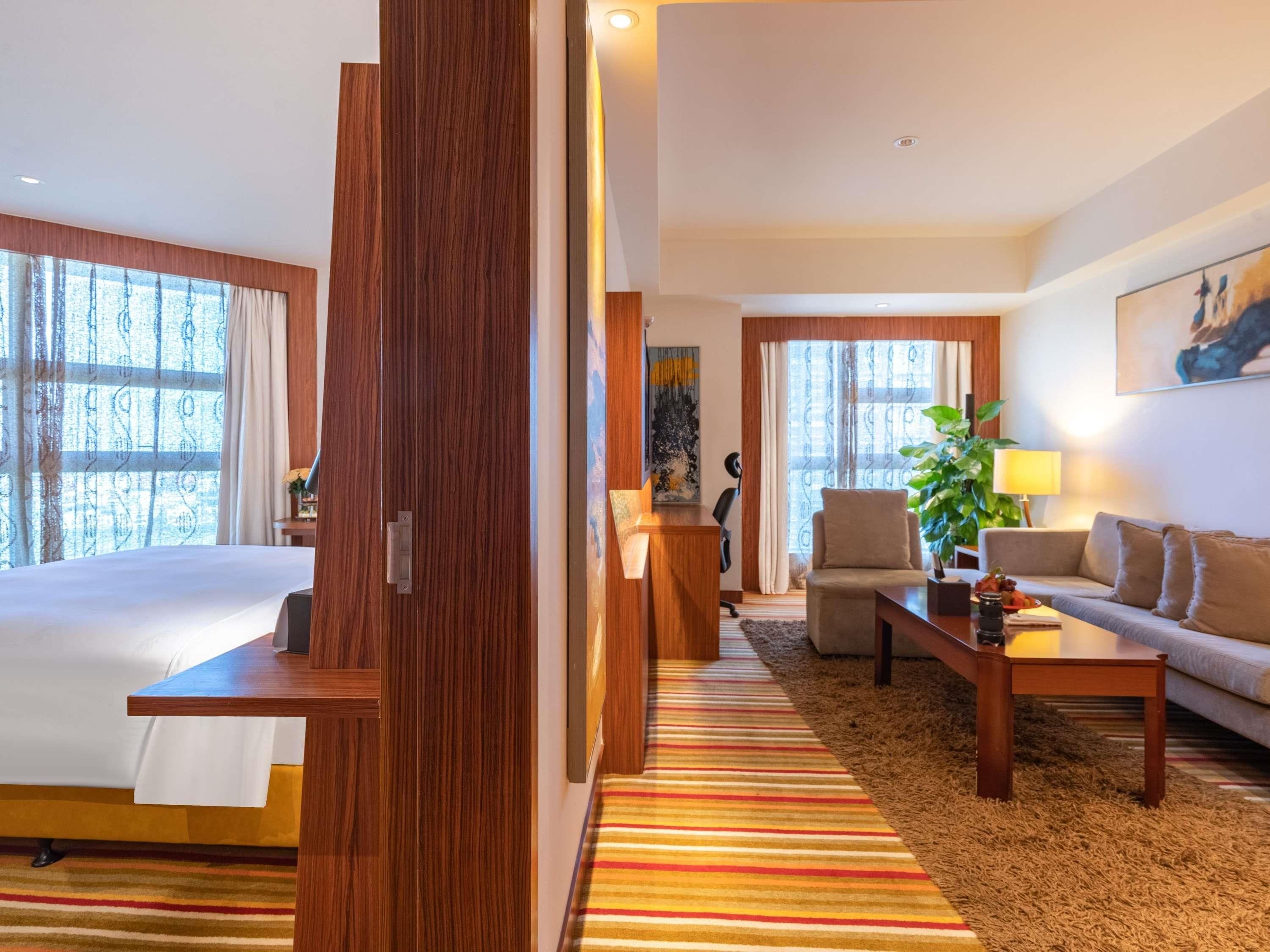 A hotel room with a bed, a desk, a sofa, a chair, a plant, and a painting on the wall.