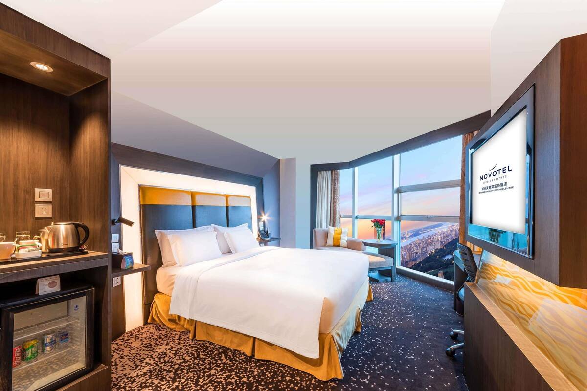 Hotelkamer met een bed, een bureau, een koelkast, een televisie en een groot raam met uitzicht op de stad.