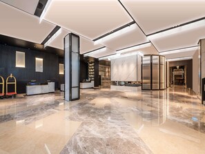 A modern hotel lobby with a reception desk, a large marble column, and a staircase leading to the upper floors.