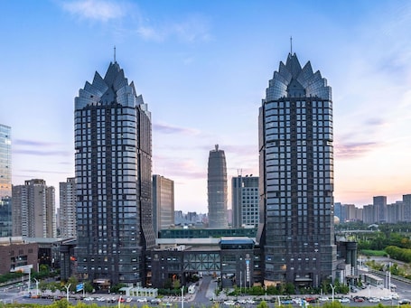 Two tall, modern buildings with a unique architectural design, surrounded by other high-rise structures in an urban setting.. Novotel Zhengzhou Convention Centre