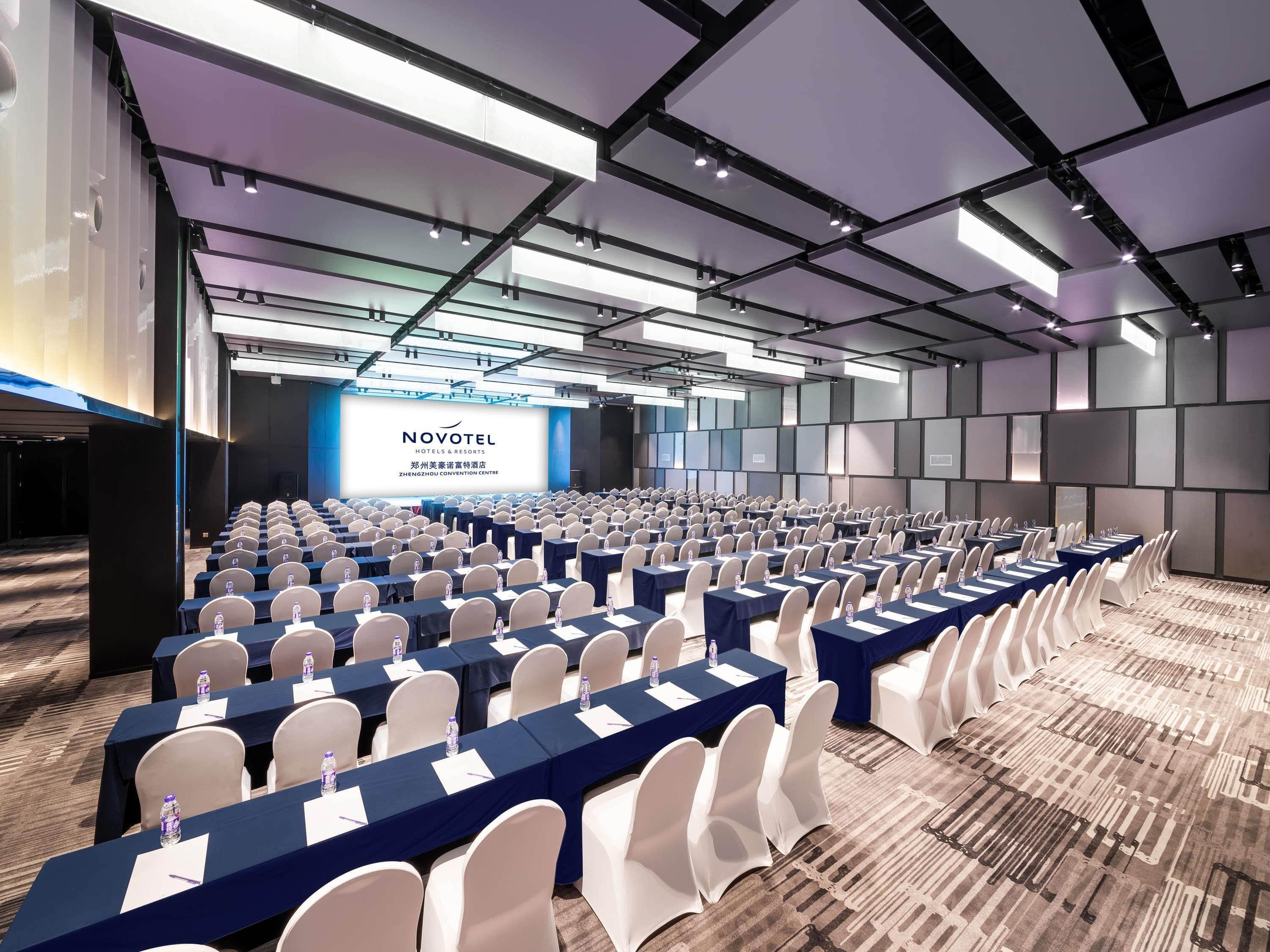 Una sala conferenze con file di tavoli e sedie, un grande schermo che mostra 'NOVOTEL HOTELS & RESORTS' e un moderno design del soffitto.