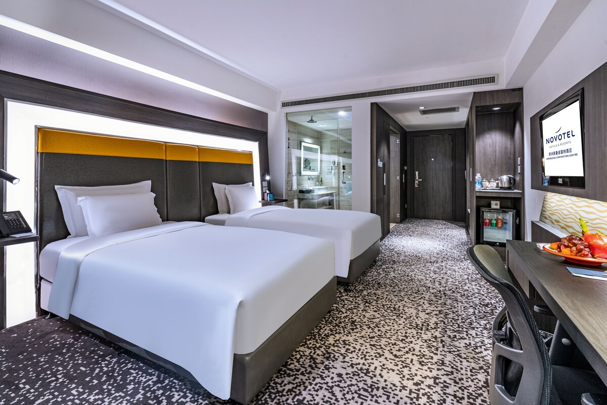 Habitación de hotel con dos camas, un escritorio con silla, zona de ducha y un televisor montado en la pared.