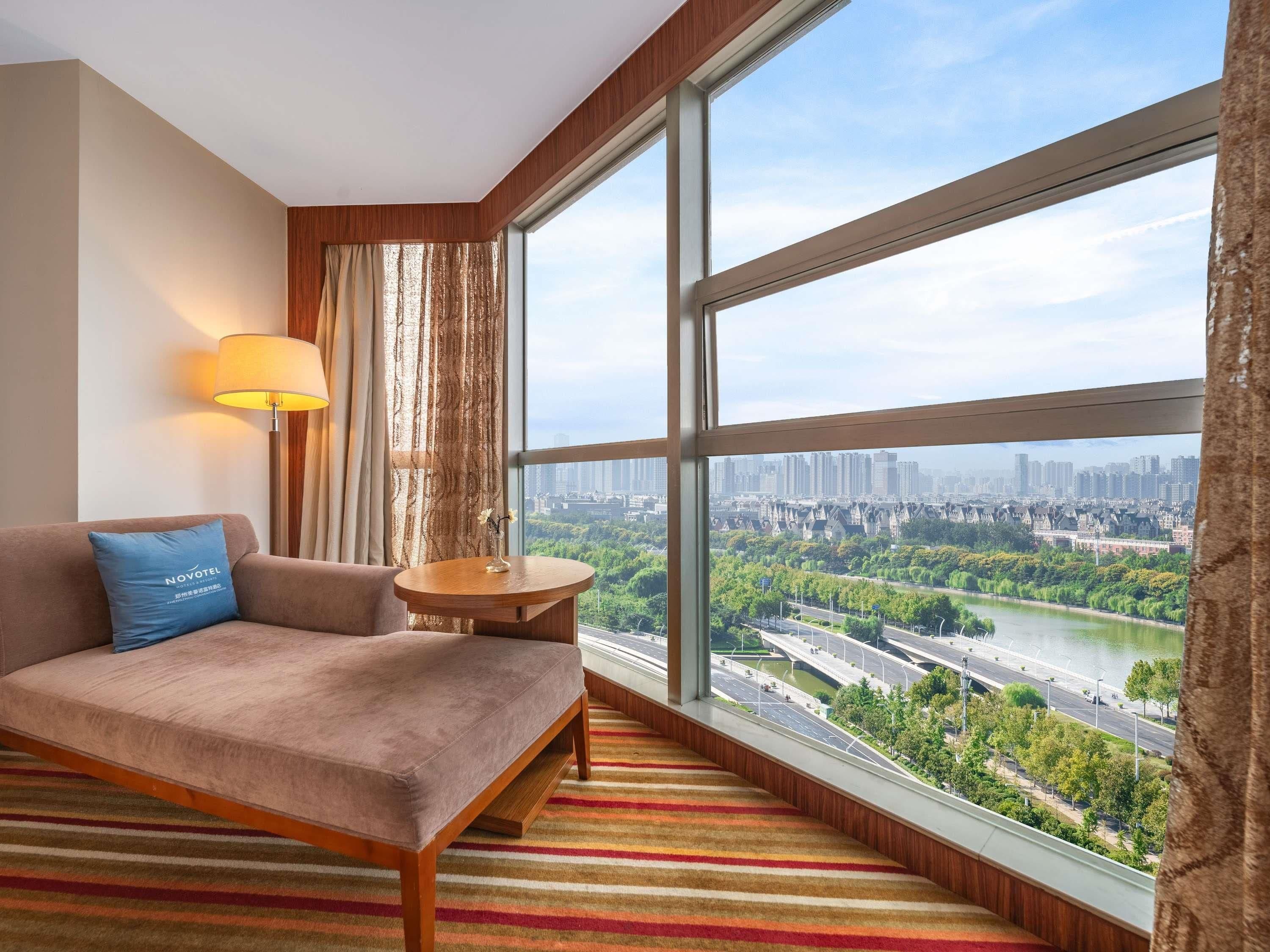 A hotel room with a large window, a sofa, a lamp, and a view of a cityscape.