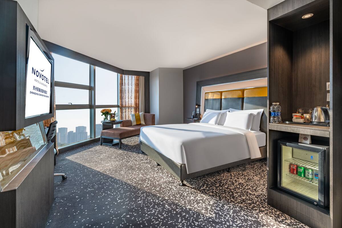 Een hotelkamer met een groot bed, een bureau met computer, een kleine koelkast en uitzicht over de stad.