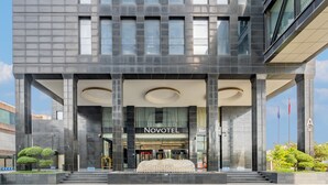 Une entrée d'hôtel moderne, avec une enseigne « NOVOTEL » bien visible, un escalier menant à l'entrée et un espace paysager agrémenté d'arbres.