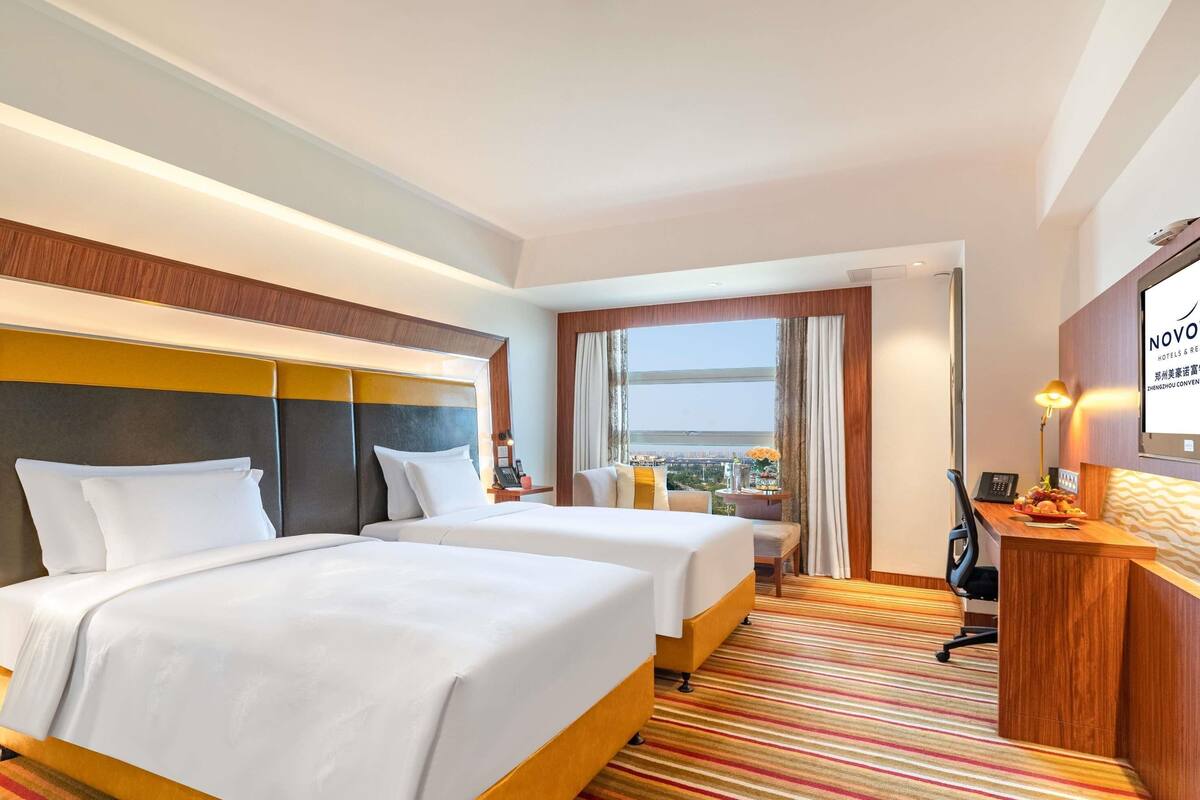 Een hotelkamer met twee bedden, een bureau, een stoel, een lamp en uitzicht op de stad.