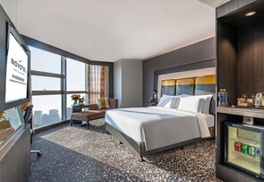 Ein Hotelzimmer mit einem großen Bett, einem Schreibtisch, einem Stuhl, einem kleinen Tisch mit einer Lampe, einer Minibar und Blick auf die Stadt.