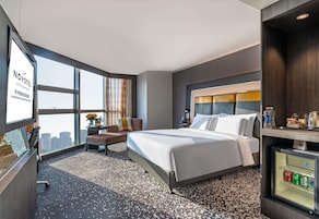 Habitación de hotel con una cama grande, un escritorio, una silla, una mesita con una lámpara, un refrigerador pequeño y vistas a la ciudad.