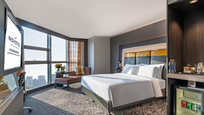 Habitación de hotel con una cama grande, un escritorio, una silla, una mesita con una lámpara, un refrigerador pequeño y vistas a la ciudad.
