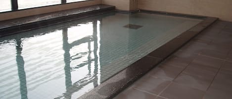 A swimming pool with clear water, tiled floor, and a window with blinds.