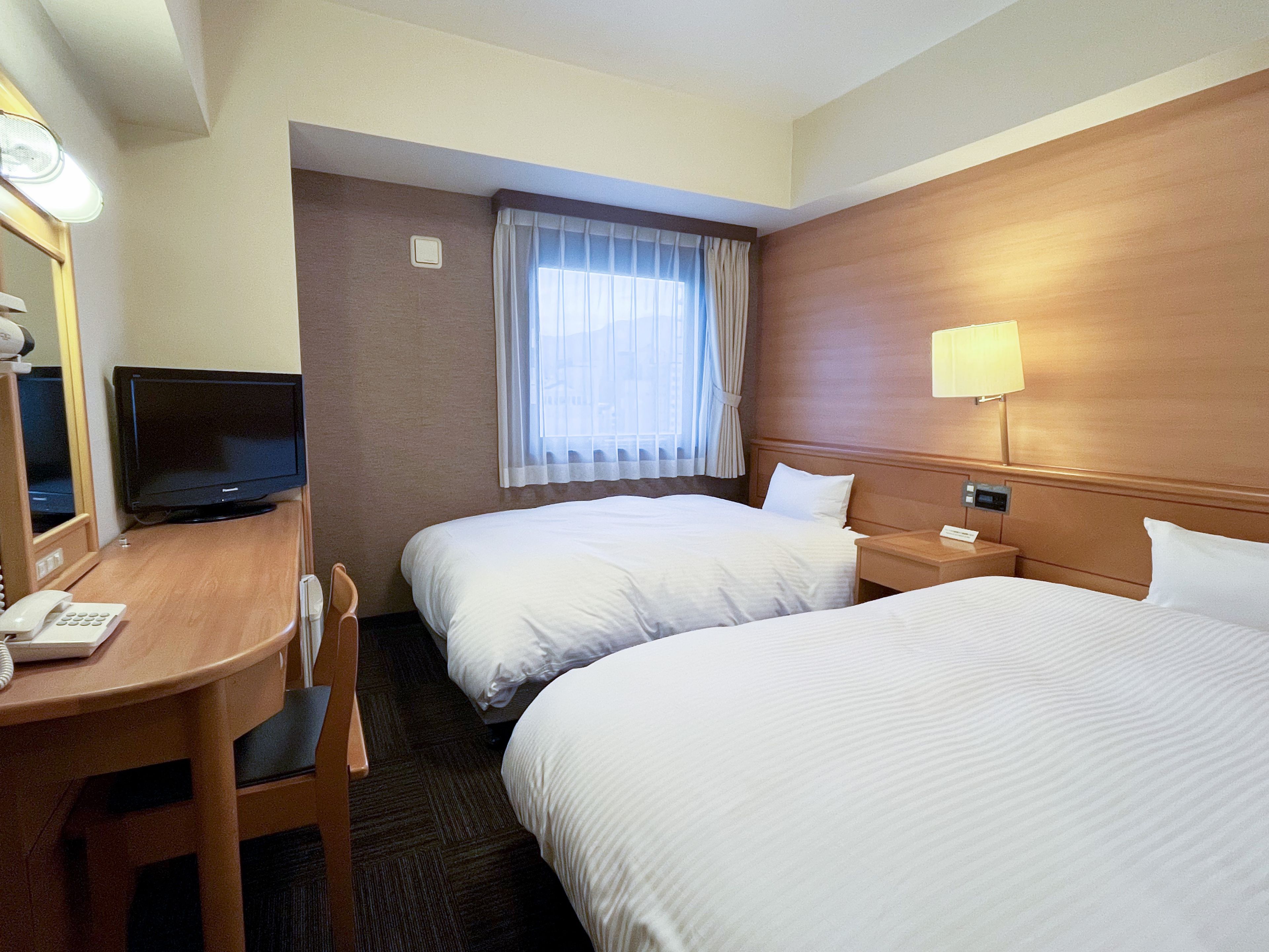 A hotel room with two beds, a desk with a telephone, a television, and a window with curtains.