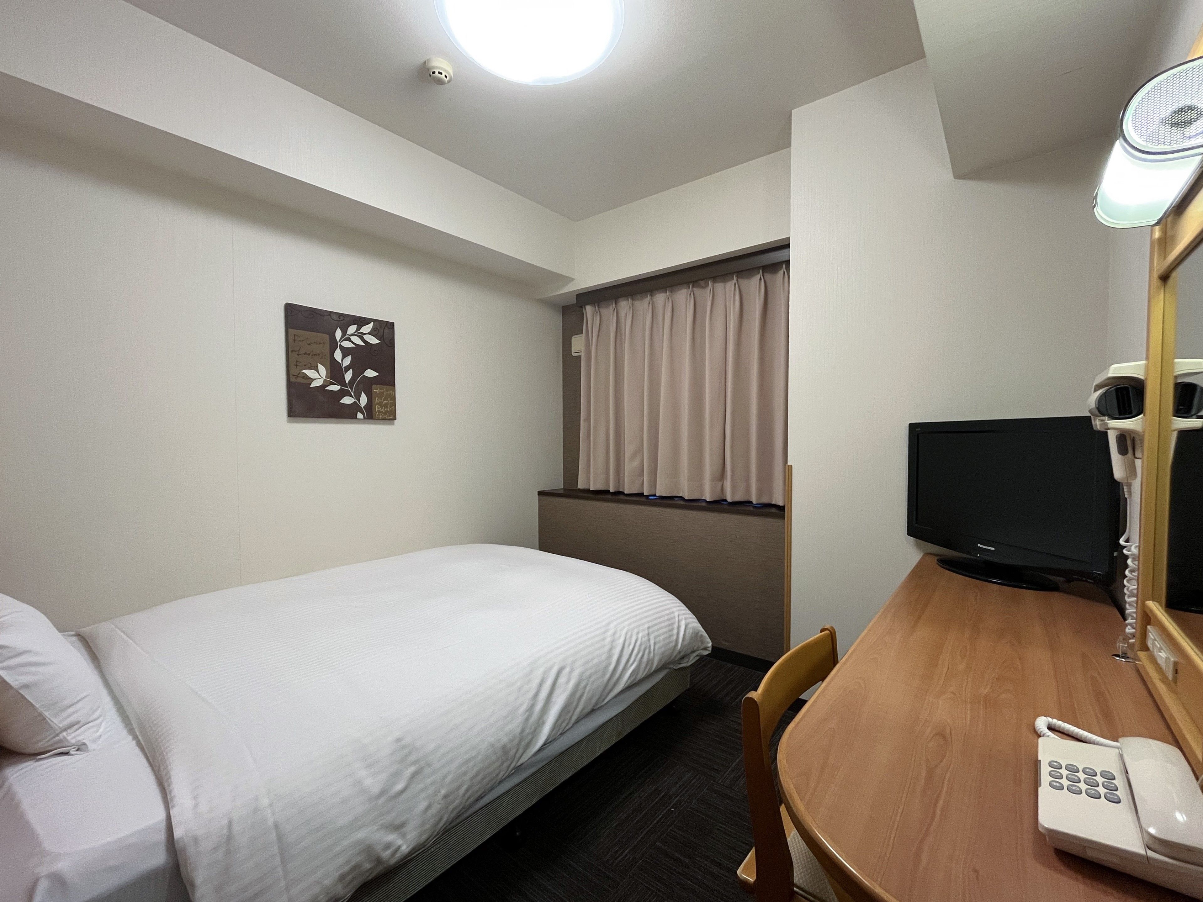Ein Hotelzimmer mit einem Bett, einem Schreibtisch mit Telefon, einem Fernseher und einem Fenster mit Vorhängen.