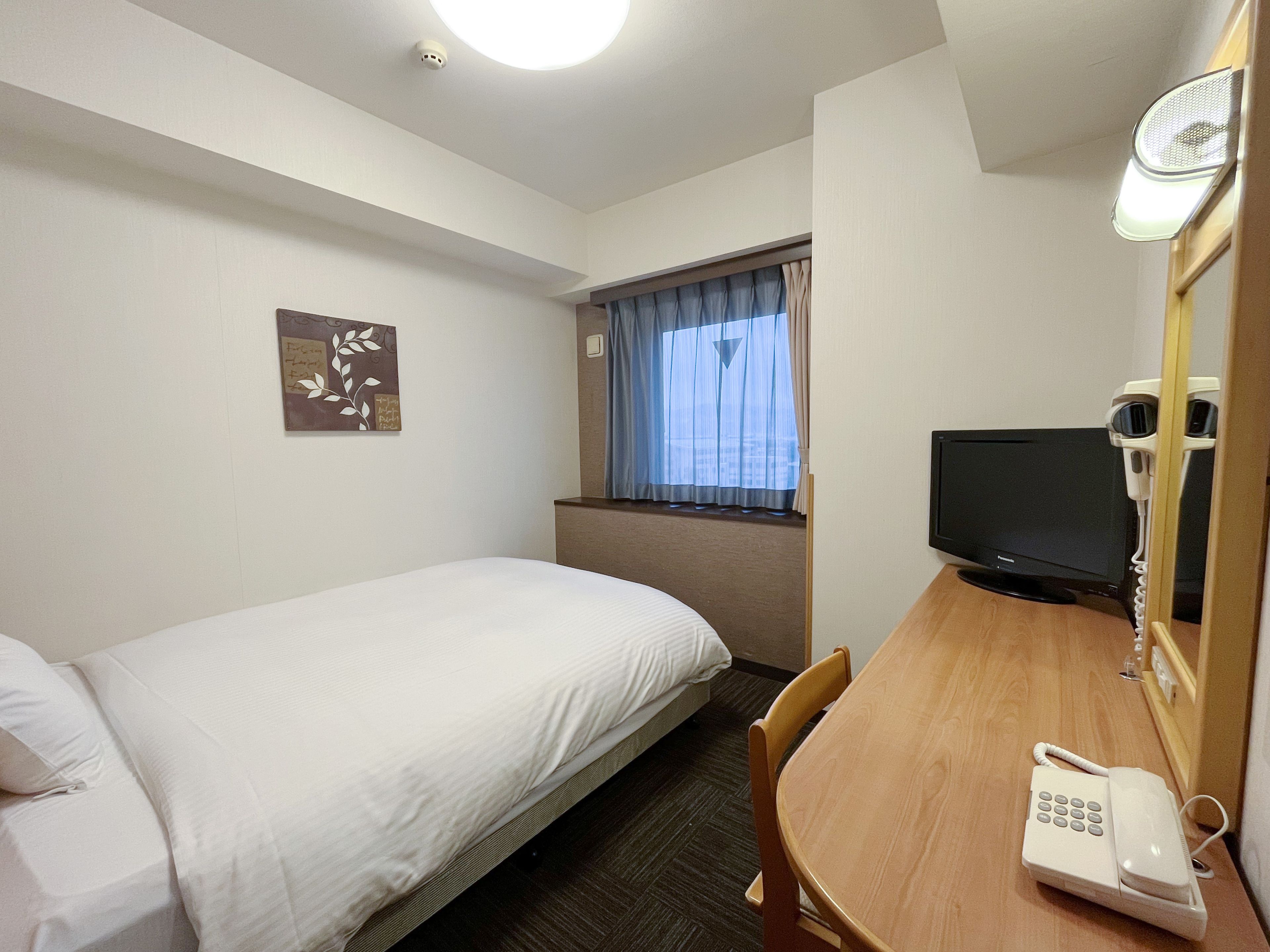 A hotel room with a single bed, a desk with a telephone, a television, and a window with curtains.
