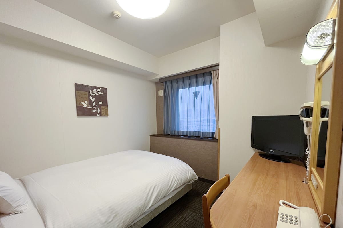 A hotel room with a single bed, a desk with a telephone, a television, and a window with curtains.