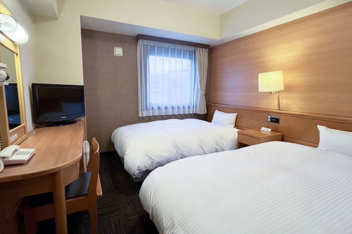 A hotel room with two beds, a desk with a telephone, a television, and a window with curtains.
