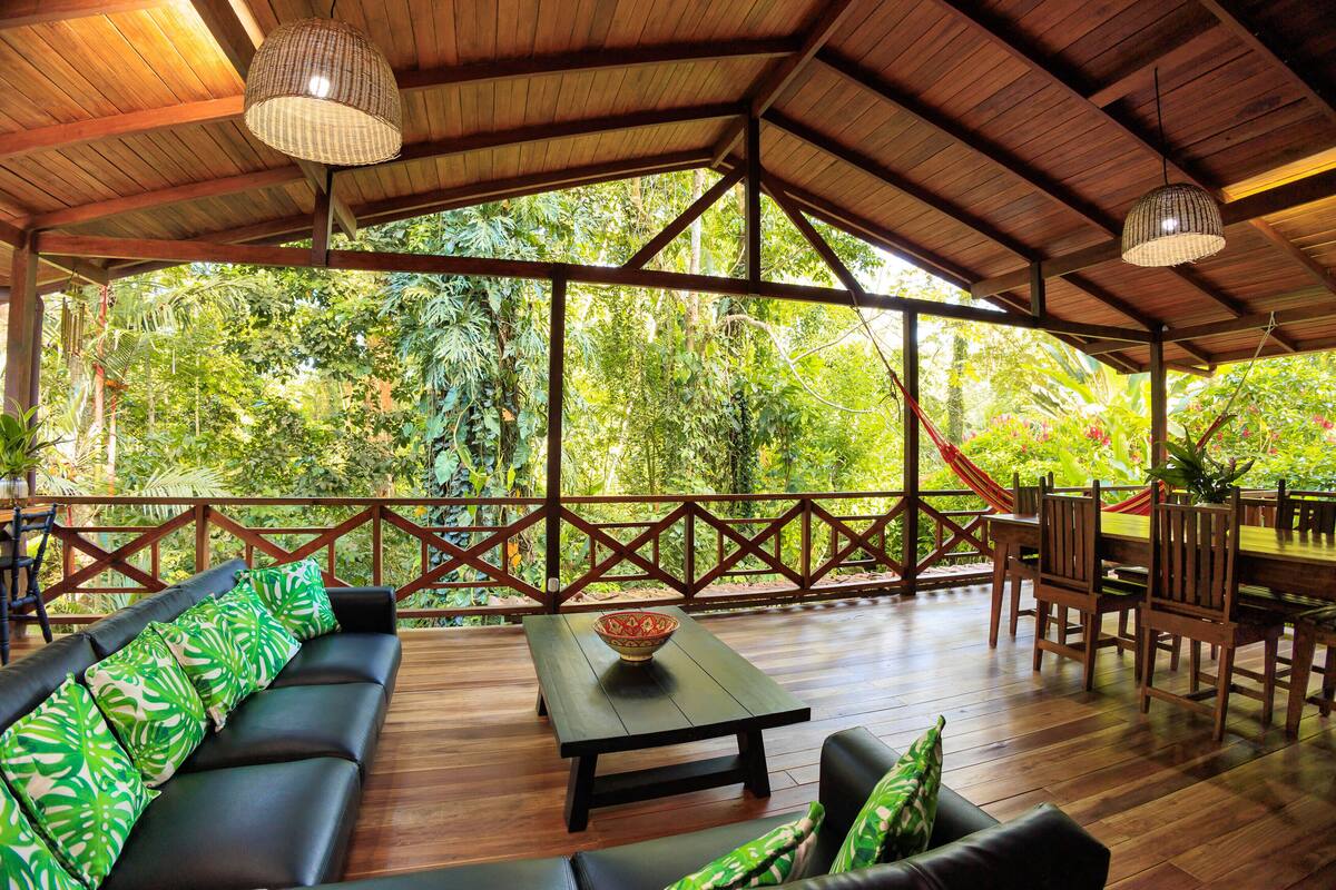 An open-air deck with a wooden floor, a black sofa with green patterned cushions, a wooden table with a bowl, and a dining area with wooden chairs and a table.