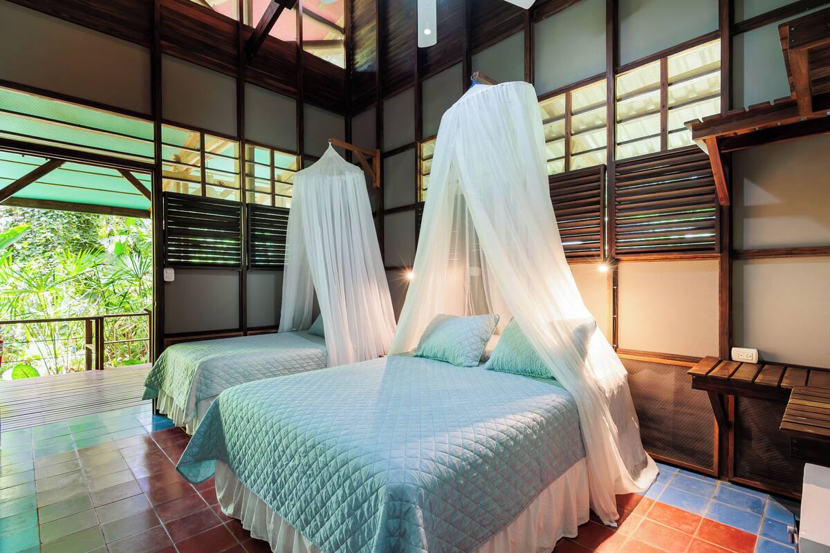 A room with two beds, each with a canopy, wooden beams, and a view of greenery outside.