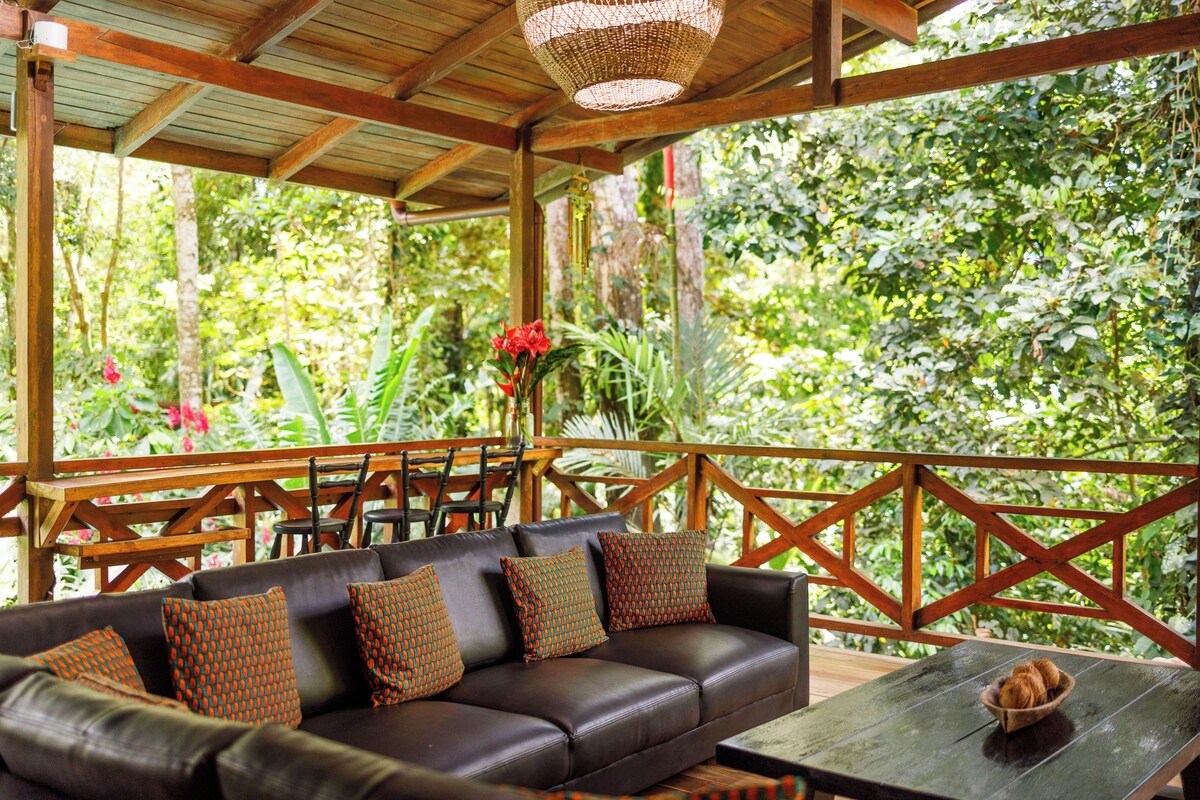 Un espace extérieur abrité, aménagé avec un banc en bois, un canapé confortable agrémenté de coussins à motifs et une table en bois rustique.