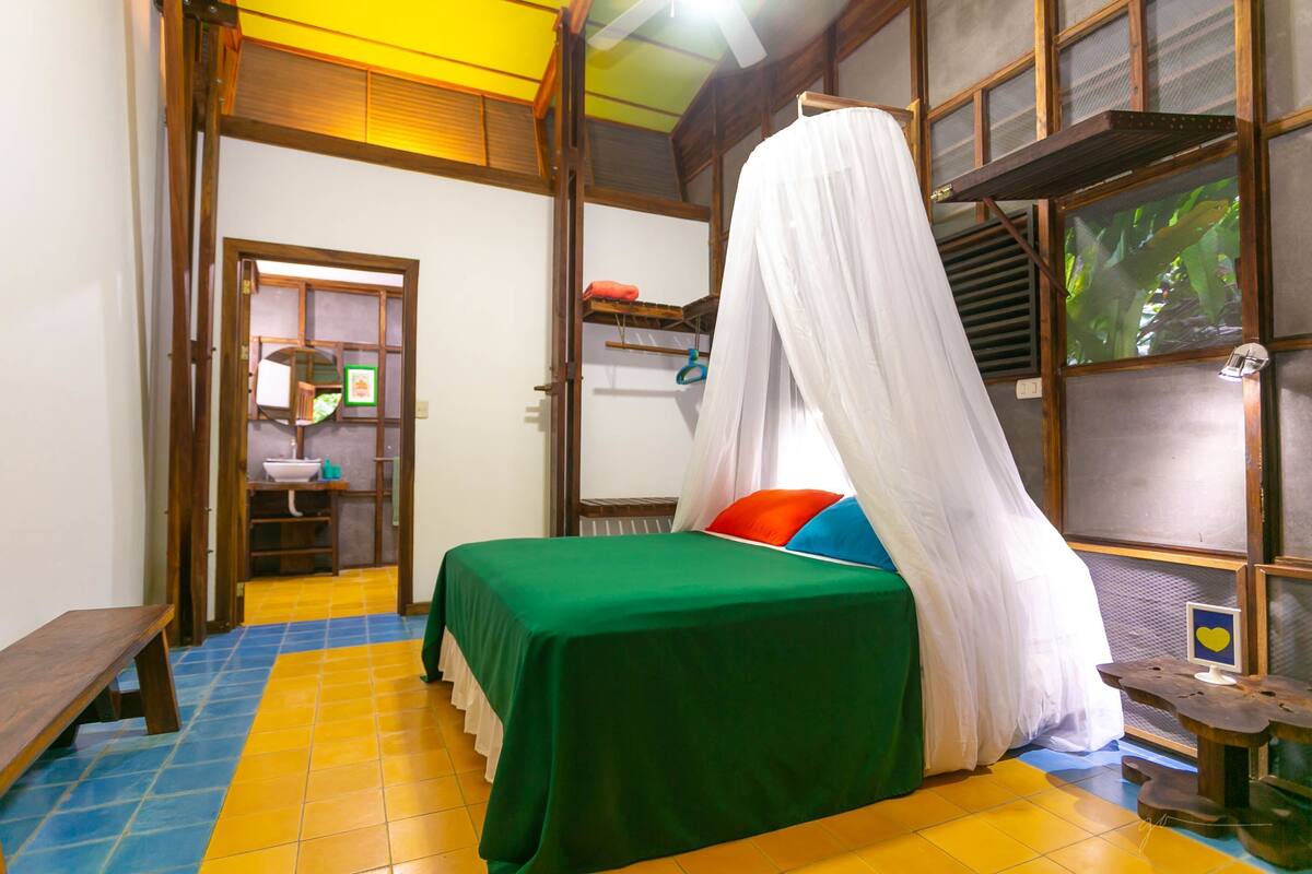 A room with a bed, a bench, a shelf with items, and a window with a view.