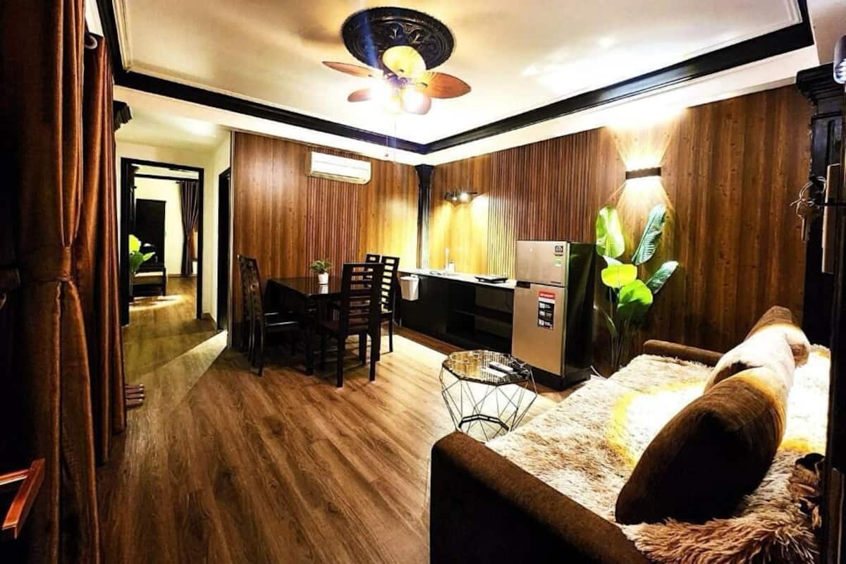 A modern living room with a sofa, a dining area with chairs, a kitchen with a refrigerator, and a ceiling fan.