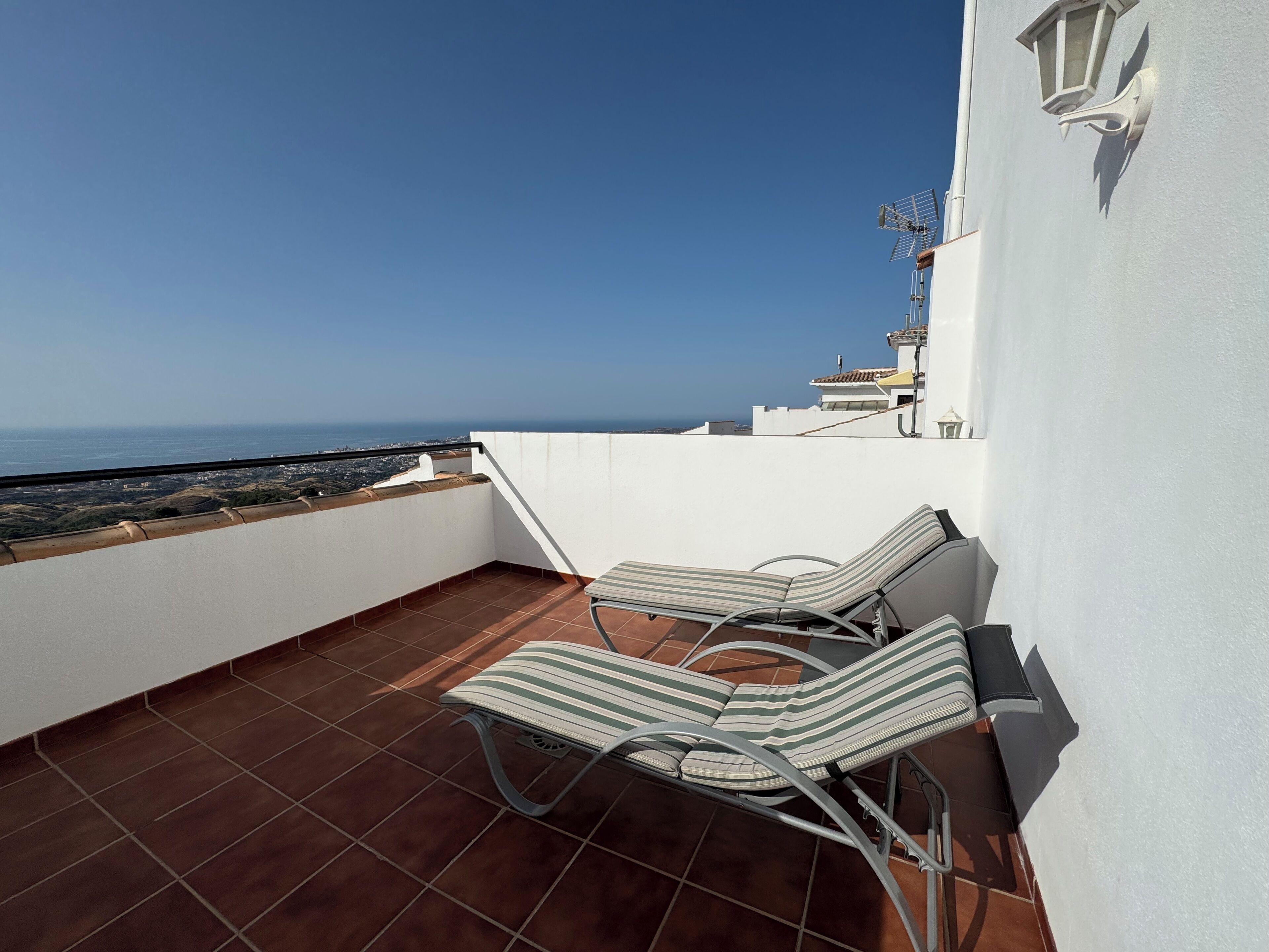 A rooftop terrace with two striped lounge chairs, a white wall, and a view of the sea.