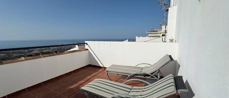 A rooftop terrace with two striped lounge chairs, a white wall, and a view of the sea.