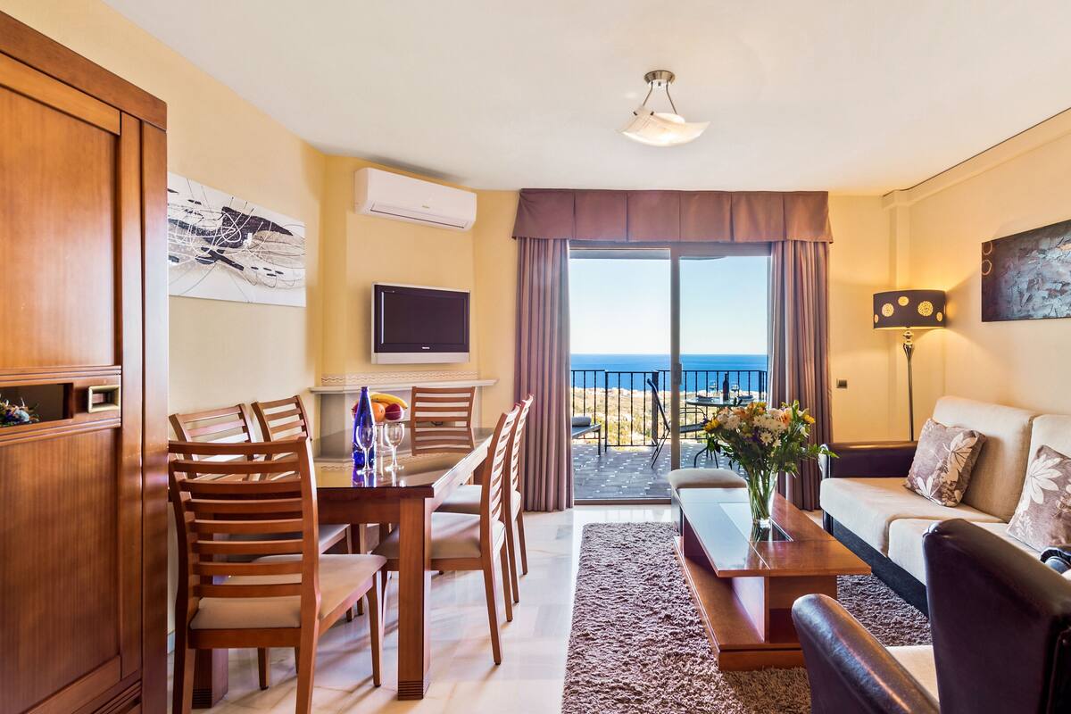 A hotel room with a dining area, a sofa, a TV, and a balcony with a view of the sea.