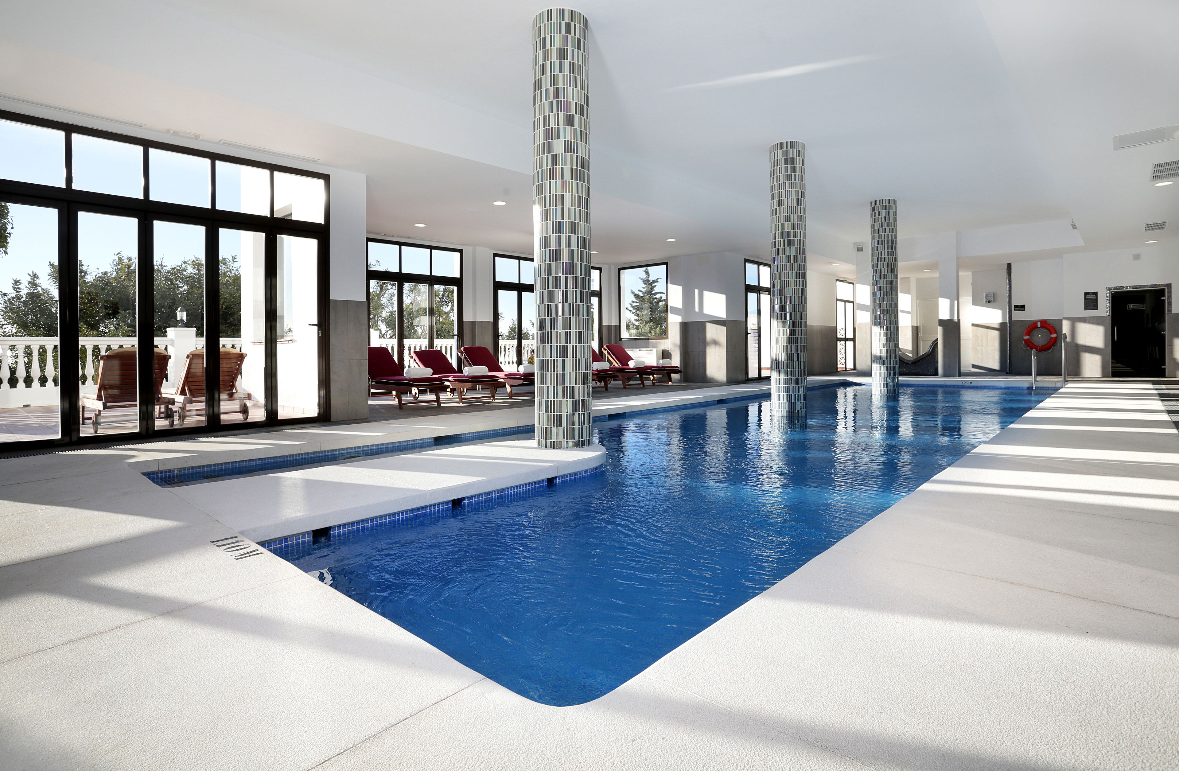 An indoor swimming pool with clear blue water, surrounded by lounge chairs and large windows.