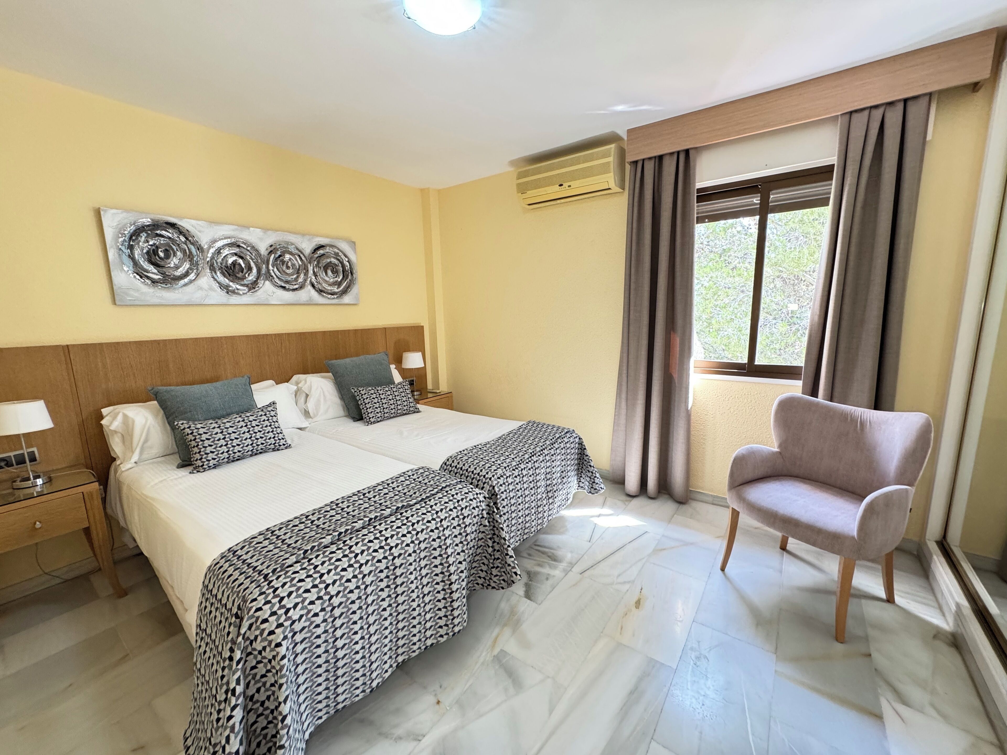 A hotel room with a double bed, two bedside tables with lamps, a chair, a large window with curtains, and a wall-mounted air conditioner.